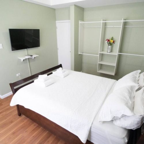 Bedroom angle featuring a wall-mounted flat-screen TV above a sleek white console shelf, open shelving unit with decorative vase, pastel green walls, and wood-framed bed.
