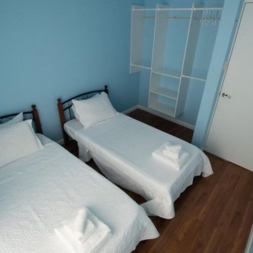 Twin bedroom featuring two single beds with crisp white linens and towels, light blue walls, built-in open closet with shelves and hanging space, and warm wooden flooring.