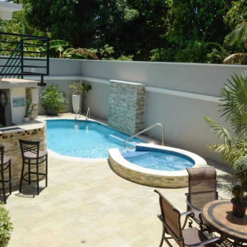Multi-purpose outdoor area with swimming pool and raised circular hot tub, shaded bar counter with stools, round dining table and chairs, and lush tropical landscaping in a private yard.