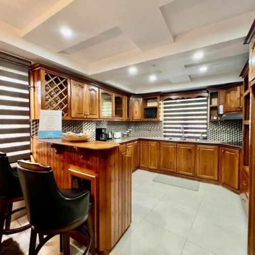 Warm L-shaped kitchen and bar area with wooden cabinetry, granite counters, built-in wine rack, mosaic tile backsplash, striped blinds and a cozy bar island with stools.