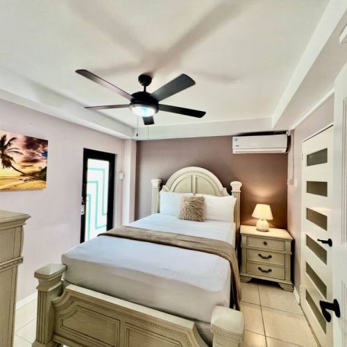 Another angle of the inviting bedroom showcasing the carved wood queen bed with crisp white linens and pillows, wall art, bedside table with lamp, ceiling fan, air-conditioning unit and modern door.