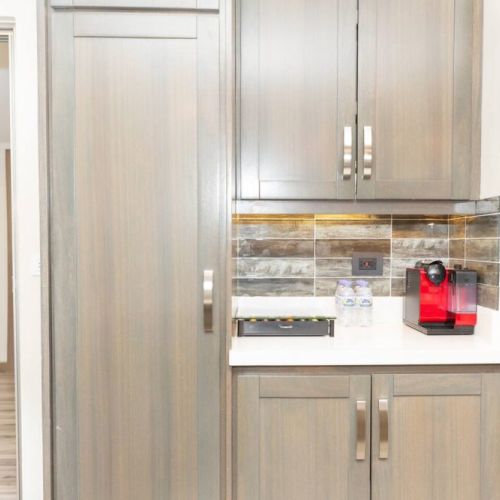 Coffee bar with coffee machine, bottled water and light wood cabinetry adjacent to kitchen