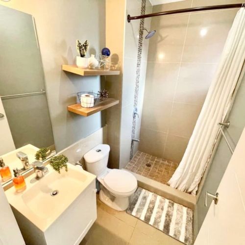 Bathroom with vanity sink, open wooden shelves with decor, toilet and walk-in shower with tiled floor, glass divider and striped rug