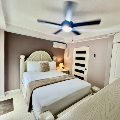 Cozy bedroom featuring a queen bed with a unique semi-circular rattan headboard, crisp white linens and beige runner with fluffy accent pillow, modern paneled door, sliding glass door with entry mat, air-conditioning unit and ceiling fan.