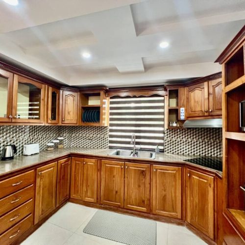 L‑shaped kitchen featuring warm wooden cabinetry with glass-front upper doors, granite countertops, mosaic tile backsplash, coffee maker, electric kettle, dish rack and stainless sink beneath modern horizontal blinds.