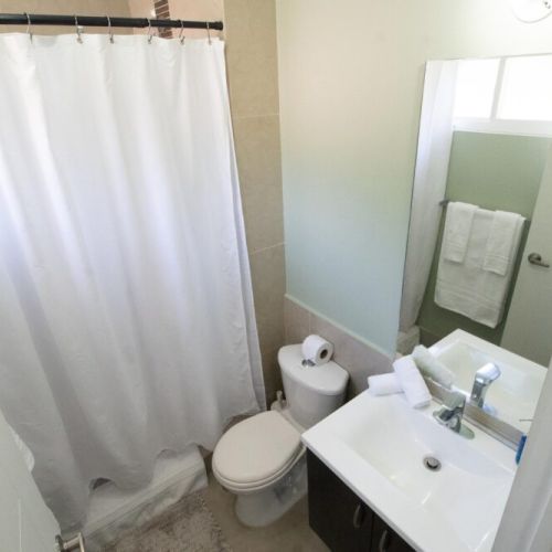 Bright bathroom featuring a combination shower with white curtain, modern vanity with sink and mirror, toilet, towel rack and window for natural light.