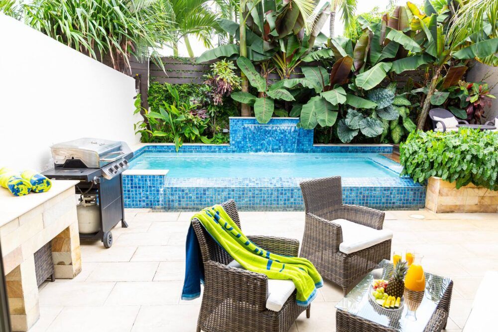 Poolside terrace with water feature, barbecue grill and wicker seating set, surrounded by lush foliage