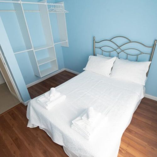 Cozy bedroom with metal-framed double bed dressed in crisp white linens and towels, light blue accent walls, warm wood flooring and built-in open closet with shelves and hanging space.