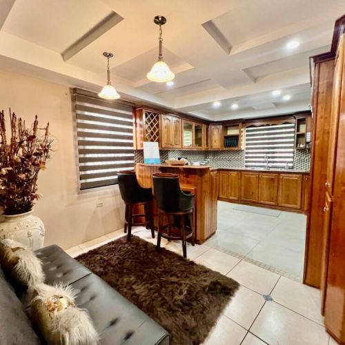 Charming lounge and bar area featuring a plush brown leather sofa with fluffy accent pillows, shaggy area rug, elegant pendant lighting, and a wooden bar with cushioned black bar stools, backed by rich cabinetry with wine storage and modern striped b