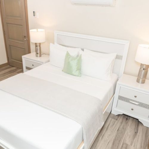 Bedroom with queen bed, white headboard, bedside tables with lamps, and wood door to en-suite bathroom