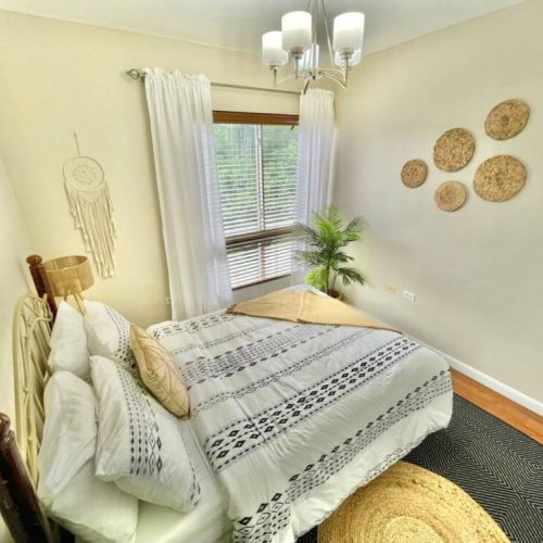 Stylish bedroom with queen bed featuring boho patterned bedding, woven wall decor, dreamcatcher, potted plant and large window with blinds