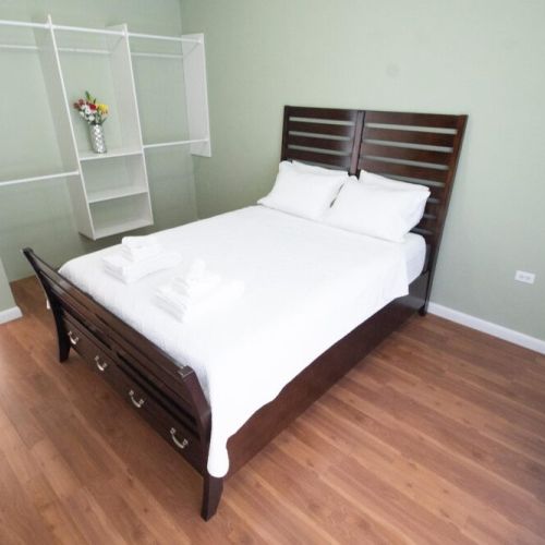 Spacious bedroom with wood-framed queen bed dressed in crisp white linens and towels, light green accent wall, built-in shelves with decor and warm hardwood flooring.