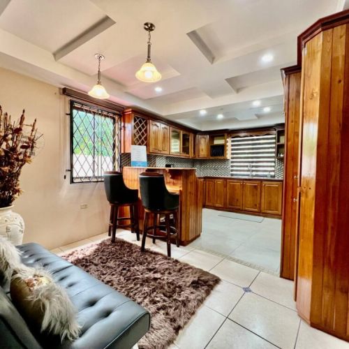Open-concept lounge and kitchen area featuring a tufted sofa with rug and decorative vase, bar table with two cushioned stools, and warm wooden L-shaped kitchen with pendant lights.