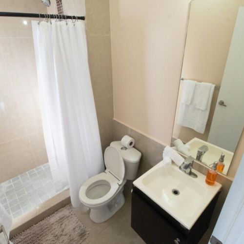 Compact bathroom featuring a tiled walk-in shower with white curtain, toilet, modern vanity with sink and mirror, and towel rack.