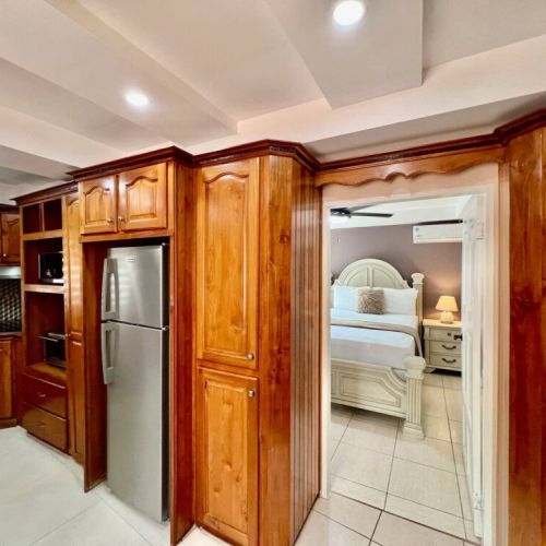 Hallway with built-in wooden pantry and storage cabinets, stainless refrigerator and doorway opening to the bedroom suite with carved wood headboard and bedside tables.