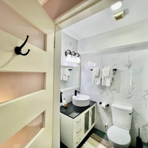 Entrance view of modern bathroom with door and black wall hook, marble-style walls, sleek white vanity with vessel sink, mirror, toilet and towel racks with neatly folded white towels.