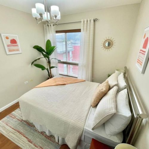 Bright bedroom with queen bed, crisp linens, accent pillows, potted plant, wall art and large window with curtains