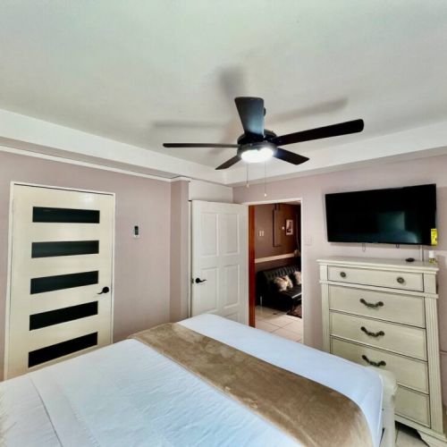 Cozy bedroom featuring a unique modern door with horizontal black stripes, white dresser with wall-mounted TV, queen bed dressed in crisp white linens with beige runner, and ceiling fan.