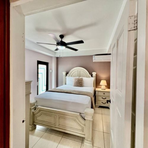 Inviting bedroom viewed from hallway showcasing a carved wood queen bed dressed in crisp white linens and pillows, nightstands with lamps, ceiling fan, air-conditioning unit and neutral decor.