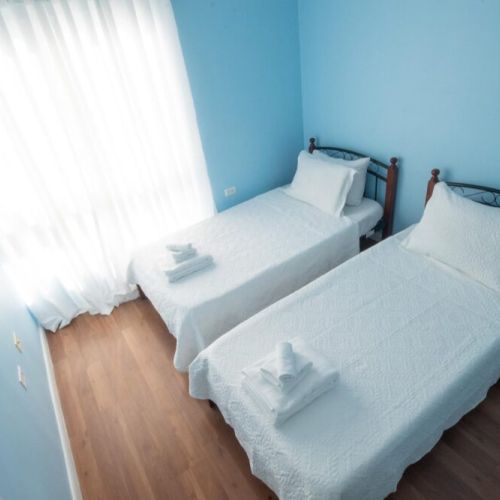 Airy twin bedroom featuring two single beds with crisp white linens and towels, light blue walls, large window with sheer curtains and hardwood flooring.