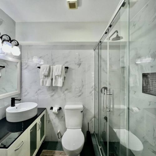 Bright modern bathroom with marble-effect walls, glass-enclosed shower with rainfall showerhead, sleek vanity with vessel sink and black countertop, large mirror and neatly folded towels.