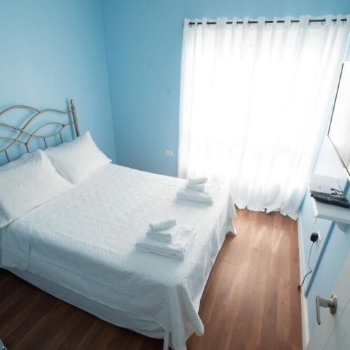 Serene blue bedroom featuring a metal-framed double bed with crisp white linens and towels, wall-mounted TV with floating shelf, large window with sheer curtains, and warm wooden flooring.
