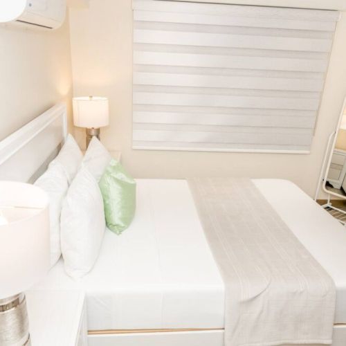 Chic bedroom with queen bed, white headboard, soft linens, green accent pillow and standing mirror near window