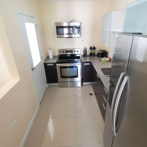 Compact galley kitchen view featuring stainless double-door refrigerator, microwave and stove, granite countertops and neutral tile flooring.
