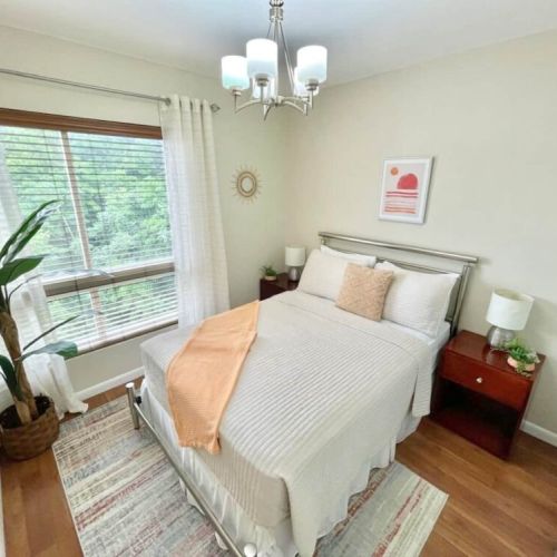 Bright bedroom with queen bed, beige bedding and orange throw, bedside tables with lamps, potted plant and large window with curtains