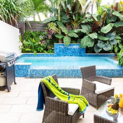 Poolside terrace with water feature, barbecue grill and wicker seating set, surrounded by lush foliage