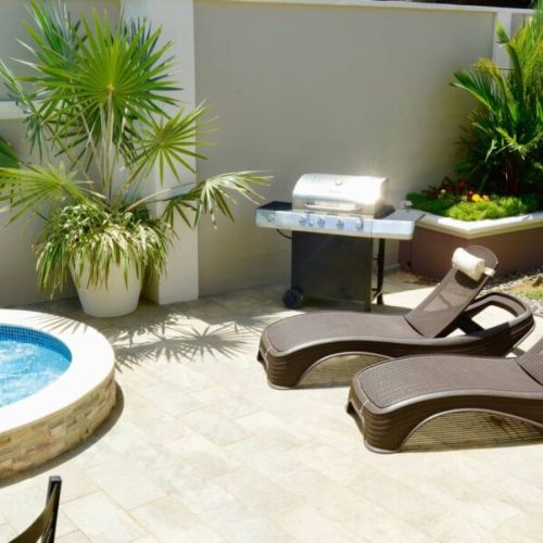 Inviting outdoor patio with circular plunge pool and hot tub, comfortable wicker lounge chairs with headrest pillows, stainless steel BBQ grill, and lush tropical landscaping.