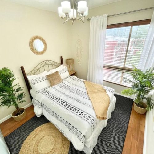 Charming bedroom with queen bed, boho patterned bedding, decorative pillows, round wall mirror, potted plants and large window with curtains