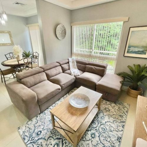 Living room with L-shaped sectional sofa, coffee table, artwork and large windows overlooking lush greenery