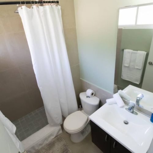 Another view of the bathroom featuring a tiled walk-in shower with white curtain, modern vanity with sink and mirror, toilet, and window bringing in natural light.