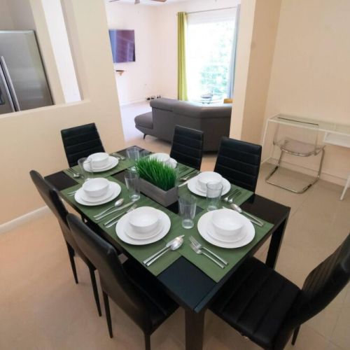 Stylish dining area featuring a black dining table set for six with green placemats and white dinnerware, sleek black chairs, and a console table with plant, open to living room.