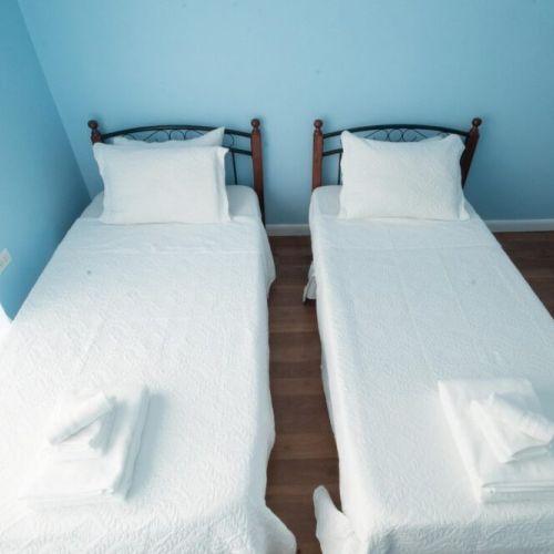 Front view of the twin bedroom showcasing two single beds with crisp white linens and towels, light blue walls, large window with sheer curtains, and warm wooden floors.