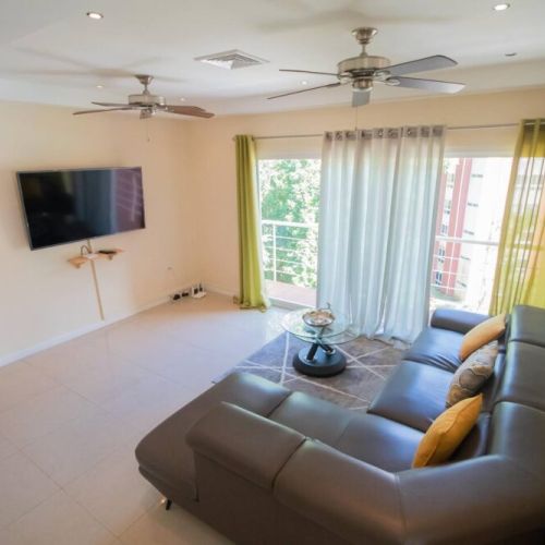 Spacious living room angle showcasing L-shaped sectional sofa, tiled flooring, large windows with sheer curtains and a wall-mounted TV.