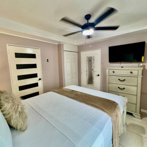 Elegant bedroom featuring a queen bed dressed in crisp white linens with a beige runner and fluffy accent pillow, modern panelled door and built-in closet, ceiling fan, wall-mounted TV and matching dresser with mirror.