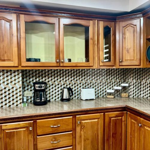 Kitchen coffee station featuring custom wooden cabinetry with glass-front upper cabinets, geometric tile backsplash, granite countertop, drip coffee maker, electric kettle and convenient outlets.