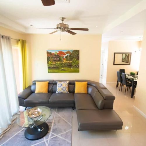 Inviting living room with sleek L-shaped sectional, glass coffee table, bright floor-to-ceiling curtains and artwork, open to dining area.