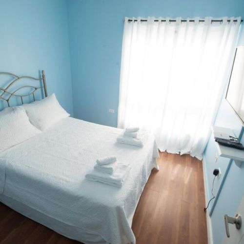 Another angle of the serene blue bedroom showcasing the metal headboard double bed dressed in crisp white linens and towels, large window with sheer curtains, wall-mounted flat-screen TV and floating shelf.