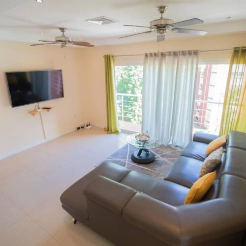 Comfortable living room with wall-mounted TV, L-shaped sectional sofa, and glass coffee table in an airy open-plan space.