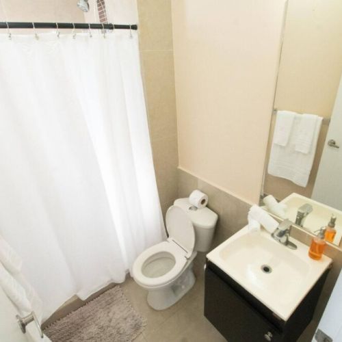 Alternate view of the bathroom showcasing the tiled walk-in shower with white curtain, small vanity with sink and mirror, toilet and bath mat.