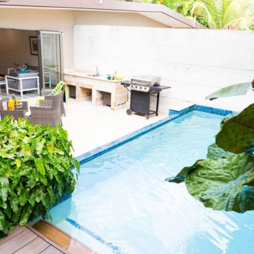 Swimming pool alongside patio  and barbecue grill, framed by large plants