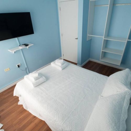 Alternate angle of the serene blue bedroom featuring a metal-framed double bed dressed in crisp white linens and towels, wall-mounted flat-screen TV, light blue walls, built-in open closet and warm wooden flooring.