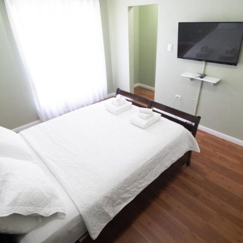 Bright bedroom with comfortable queen bed dressed in crisp white linens and towels, warm wood flooring and wall-mounted TV with shelf.